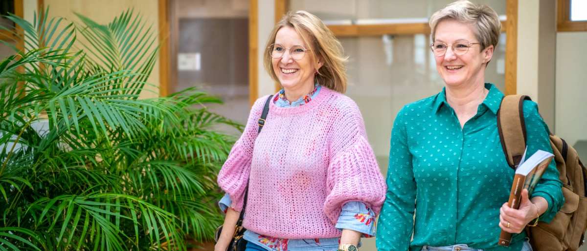 Twee studenten lopen naast elkaar en lachen, terwijl ze door de gang lopen.