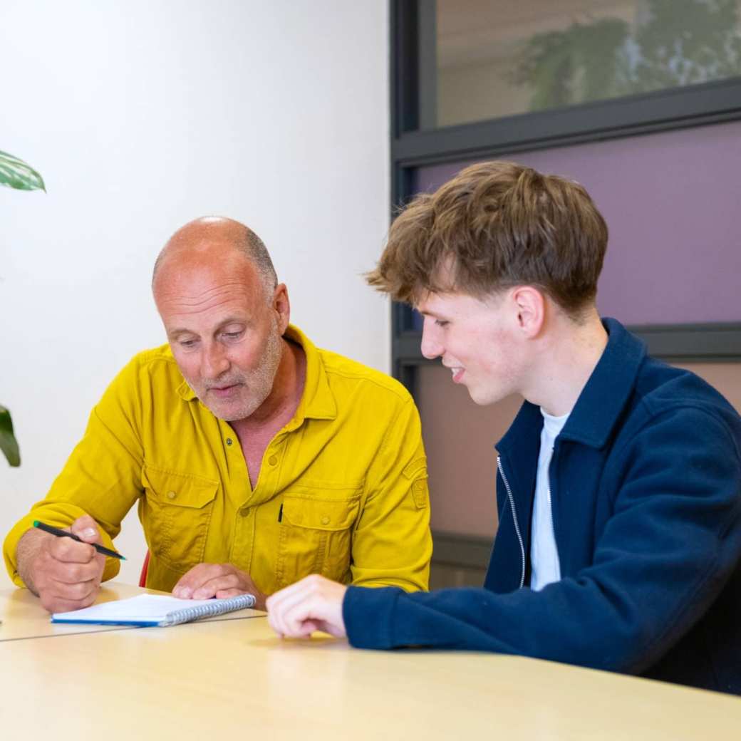 Docent en student zitten aan een tafel in een overlegruimte en bespreken studievoortgang.