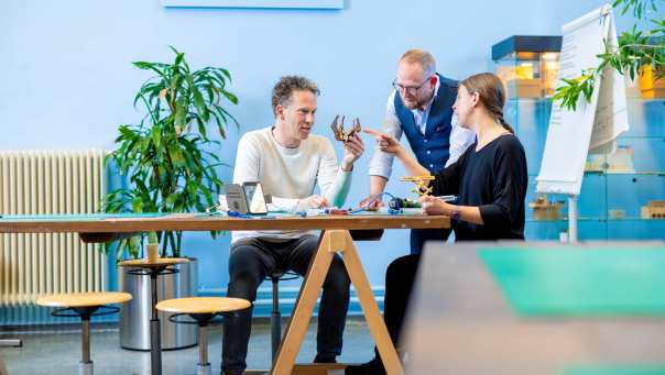 Drie studenten hebben overleg aan de tafel bij een blauwe muur