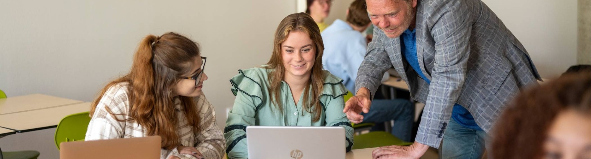 Twee studenten in de klas aan tafel en overleggen met docent die uitleg geeft.