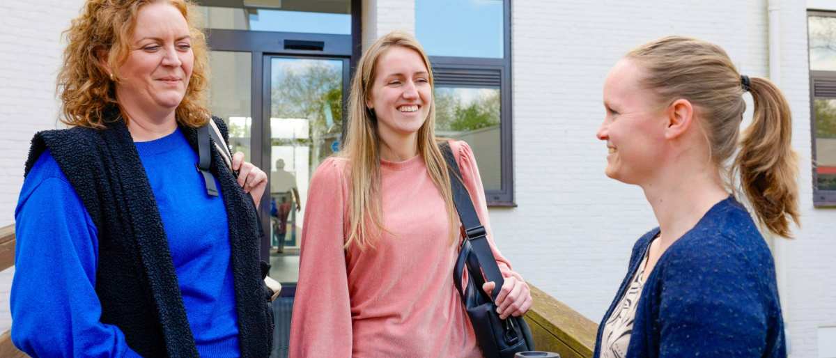 drie vrouwen staan samen buiten, in gesprek. Roze trui is aan het lachen.
