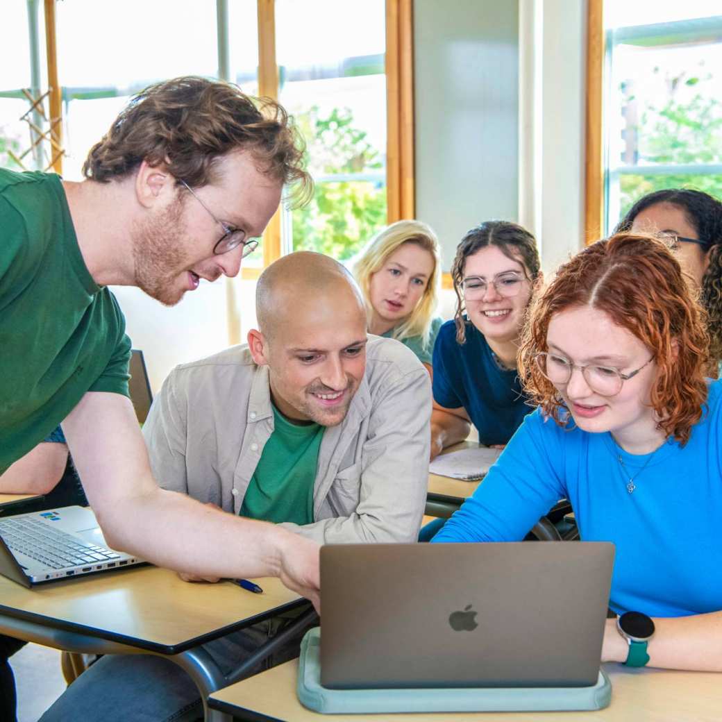 Docent wijst iets aan op een laptop, studenten kijken mee