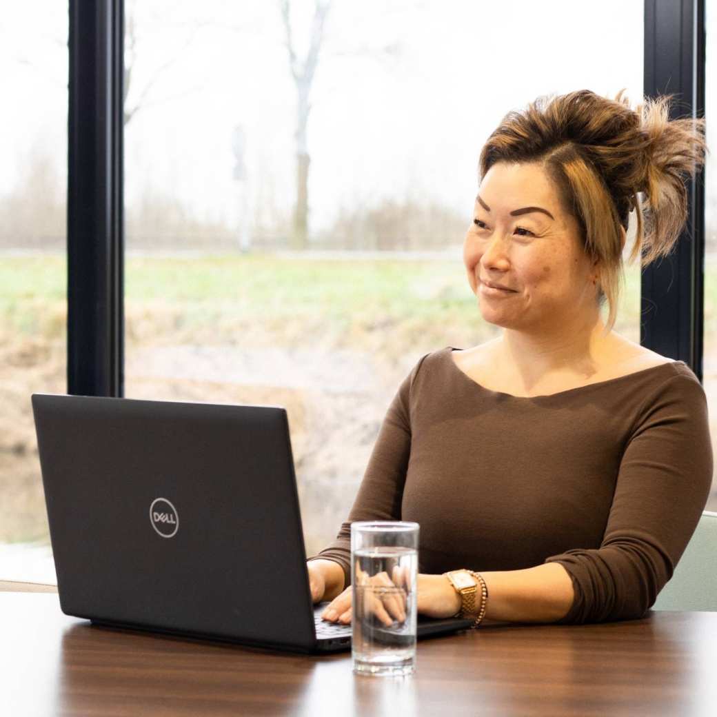 Dame poseert met een glimlach, achter een laptop. Een glas water staat naast haar op het bureau.