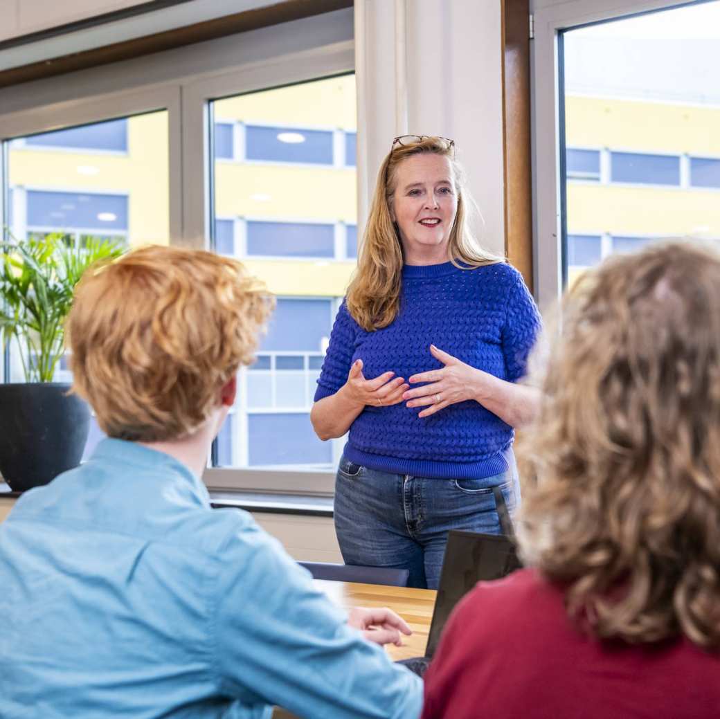 Docent in blauw shirt geeft les aan 3 studenten