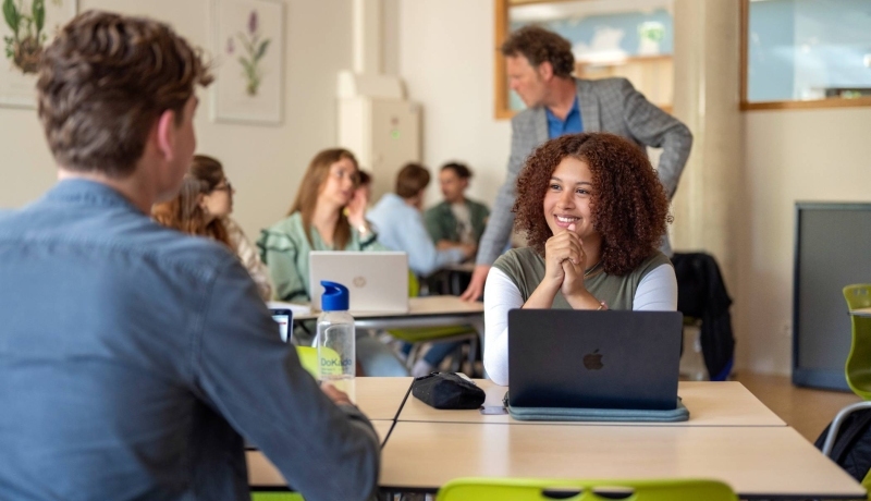 Meisje lacht en overlegt met een jongen, in de klas aan tafel. 