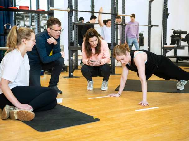 Student staat in een plank positie, 3 studenten kijken toe. 