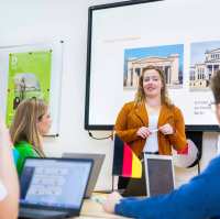 Docent geeft aan twee studenten uitleg en wijst daarbij naar de pagina in het boek dat de studenten in de hand hebben.