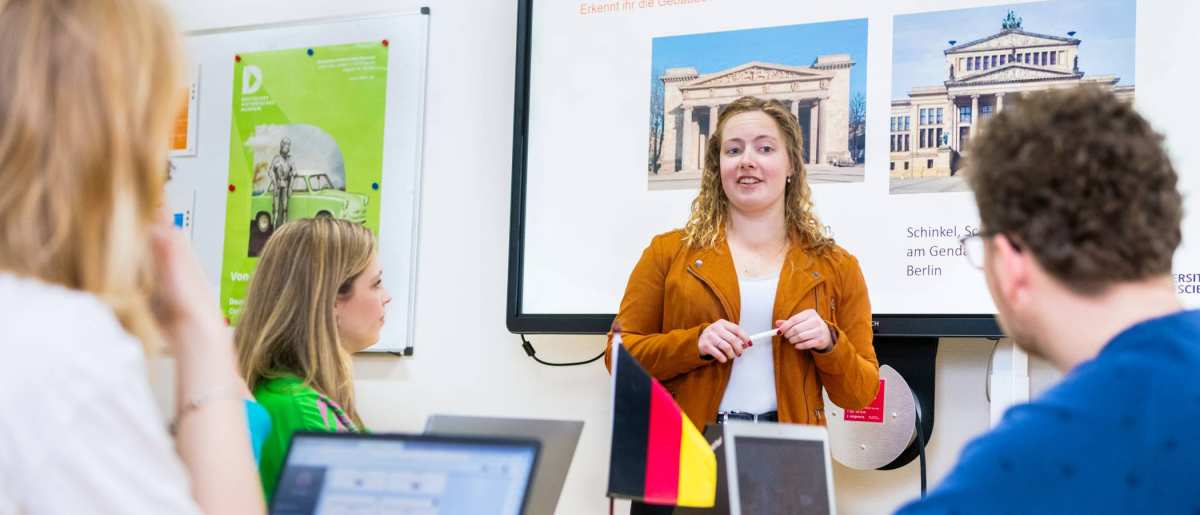 Student leraar Duits in oranje/bruin jasje geeft een lesje aan haar medestudenten. Ze staat voor een PowerPoint.

