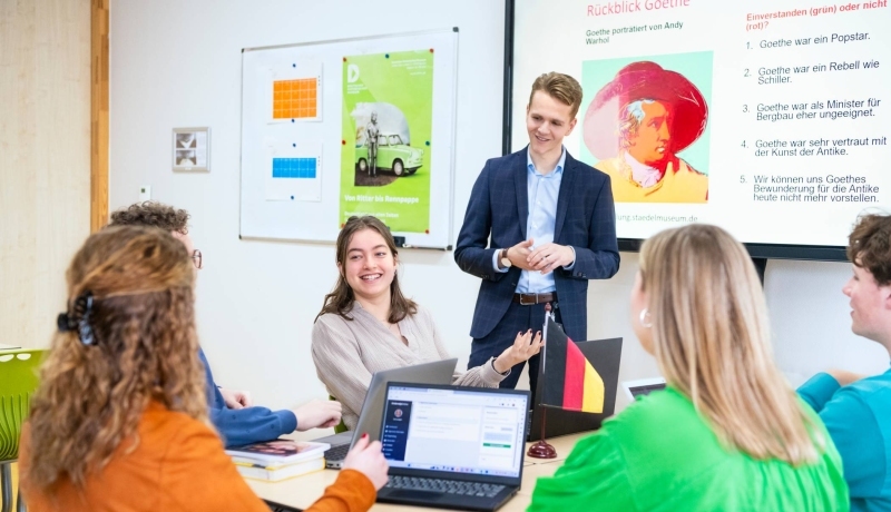 Studenten zitten samen aan tafels met laptops en boeken in het klaslokaal. De leraar staat ervoor en praat met hen mee.