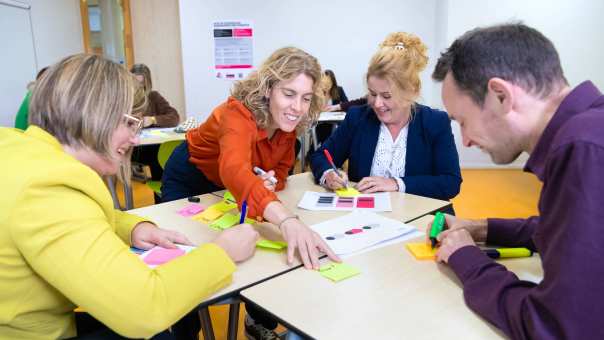 Een groepje van vier mensen in gesprek met post-its op tafel in een lokaal met anderen