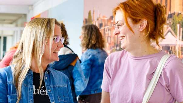 Een vrouw met een roze shirt en rood haar praat met een blonde vrouw met een spijkerjasje aan op de gang