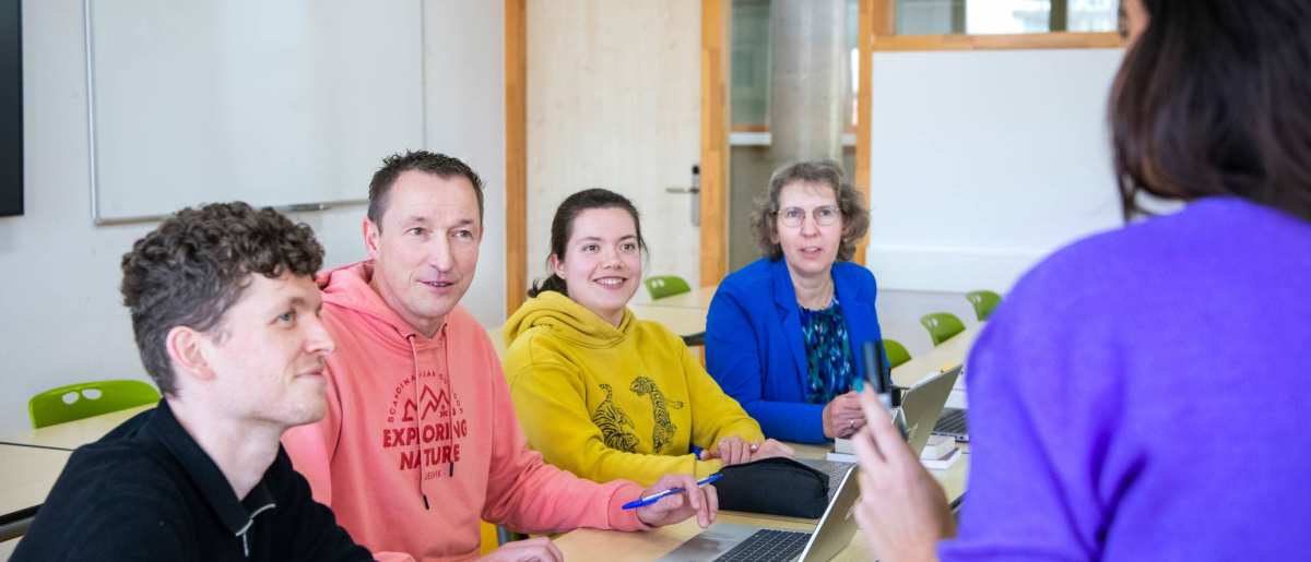 Foto Master Academie Educatie. 4 masterstudenten zitten achter een tafel en luisteren naar een mevrouw die iets vertelt. 