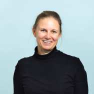 Portrait headshot. The picture is taken in front of a blue background and the subject is wearing a a black turtleneck shirt. Her hair is brunette and tied behind her head. Her eyes are blue. Her right hand is in the right pocket of her beige and brown plaid pleated pants.. She is facing the camera, with shoulders slightly towards the left.