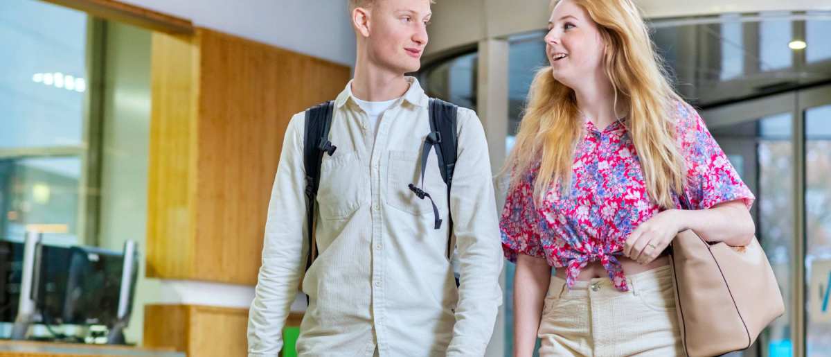 Twee studenten lopen naast elkaar door de gang en vertellen met elkaar.