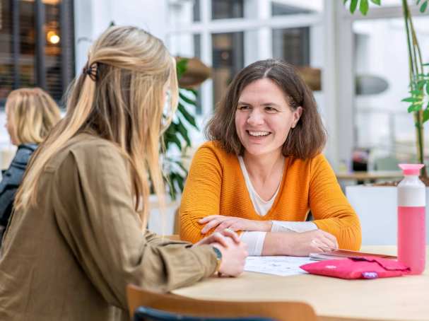 Vrouw in oranje trui lacht naar medestudent.