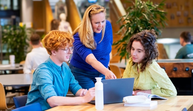 De lerares wijst naar de laptop waar twee studenten bij zitten.