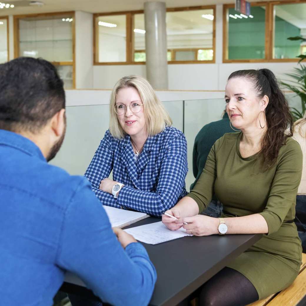 Twee studenten zitten aan een tafel en luisteren aandachtig naar de student die voor hun zit.