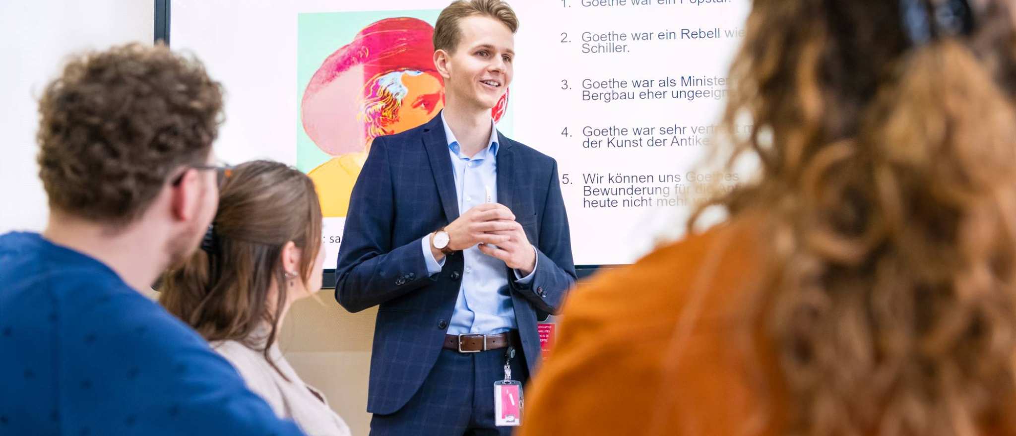 Leraar Duits in pak staat voor de klas met zijn handen in elkaar gevouwen en legt iets uit. Hij heeft een Powerpoint op de achtergrond openstaan.
