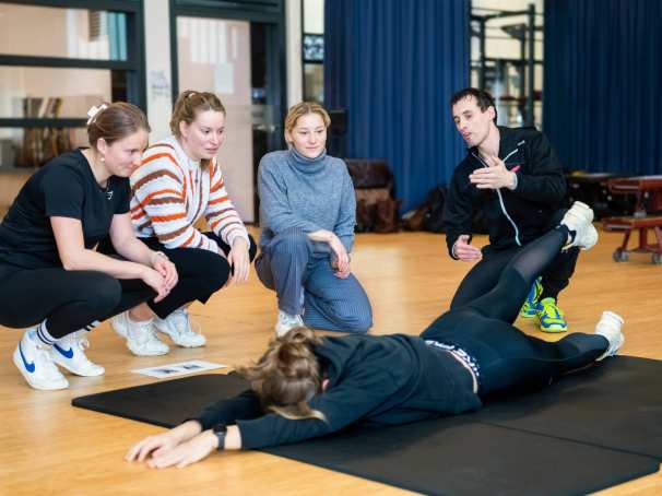 Student ligt op haar buik op de grond, heft een van haar benen de lucht in. Docent en 3 studenten kijken toe. 