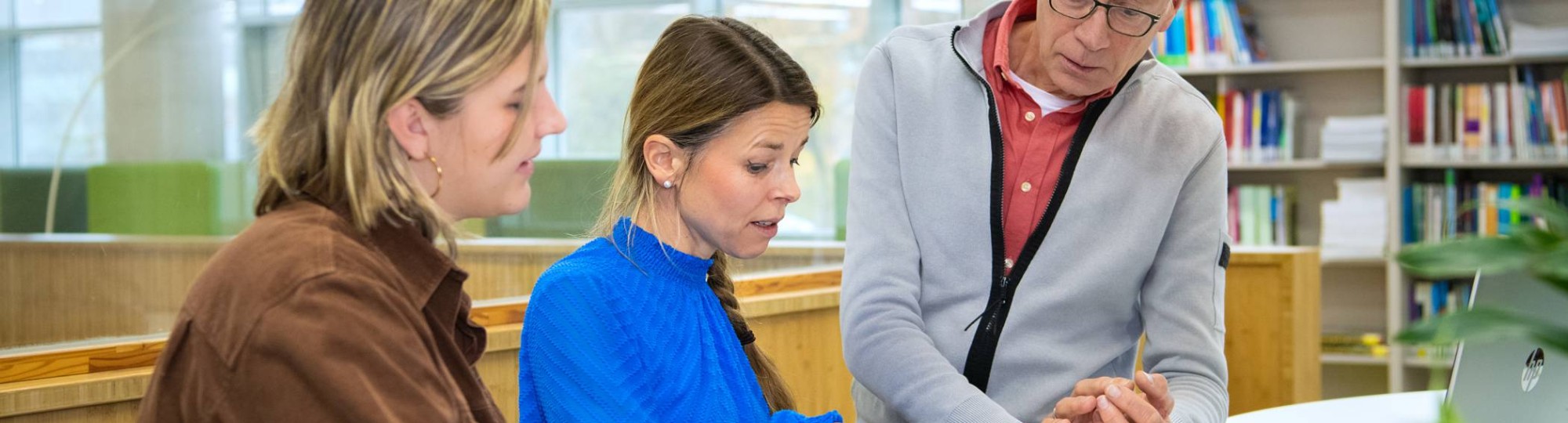 Een man en twee vrouwen in gesprek aan een tafel. Een vrouw wijst iets aan op papier.