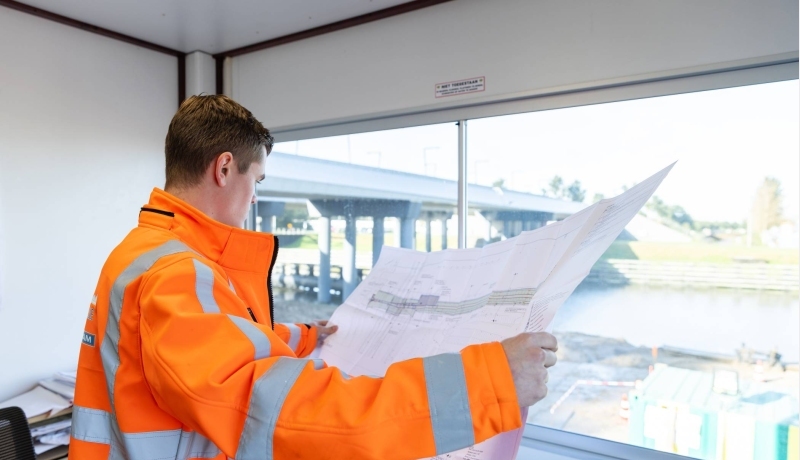 opzichter in de directiekeet kijkt met tekeningen in de hand uit over het bouwterrein