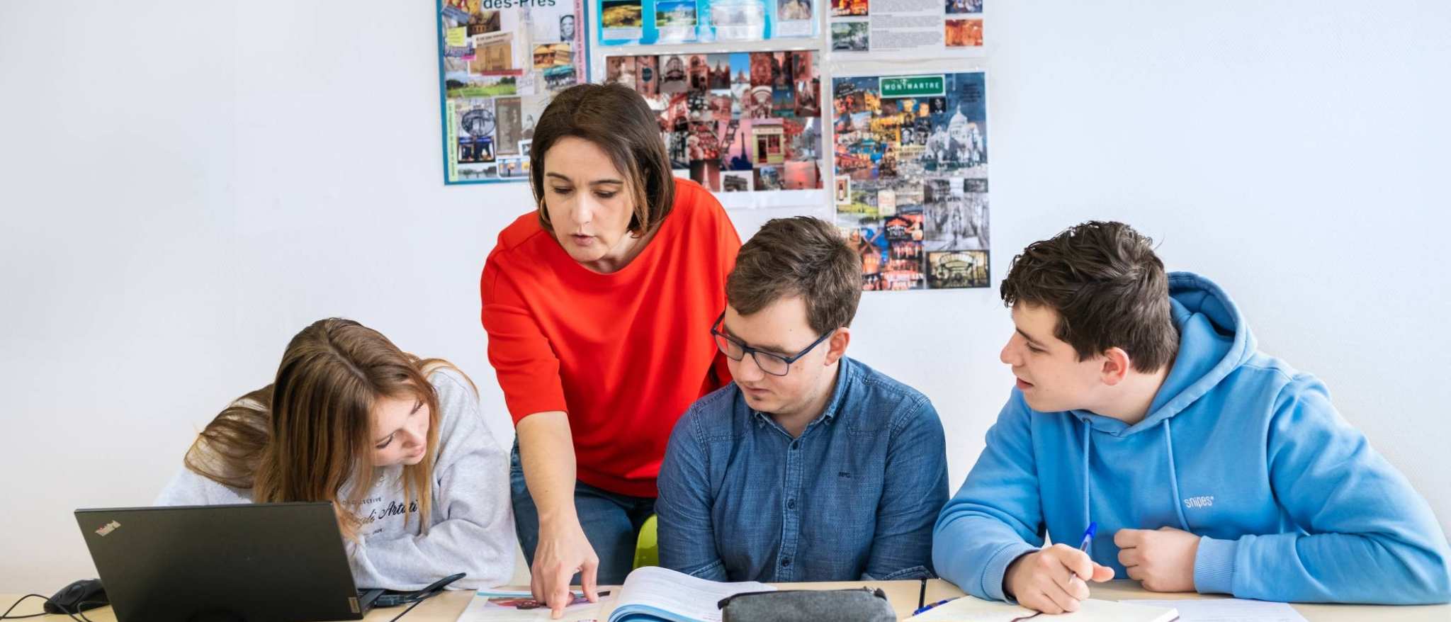 Drie studenten zitten op een rij en de docent staat achter hen. Ze wijst in het boek en de studenten kijken mee.