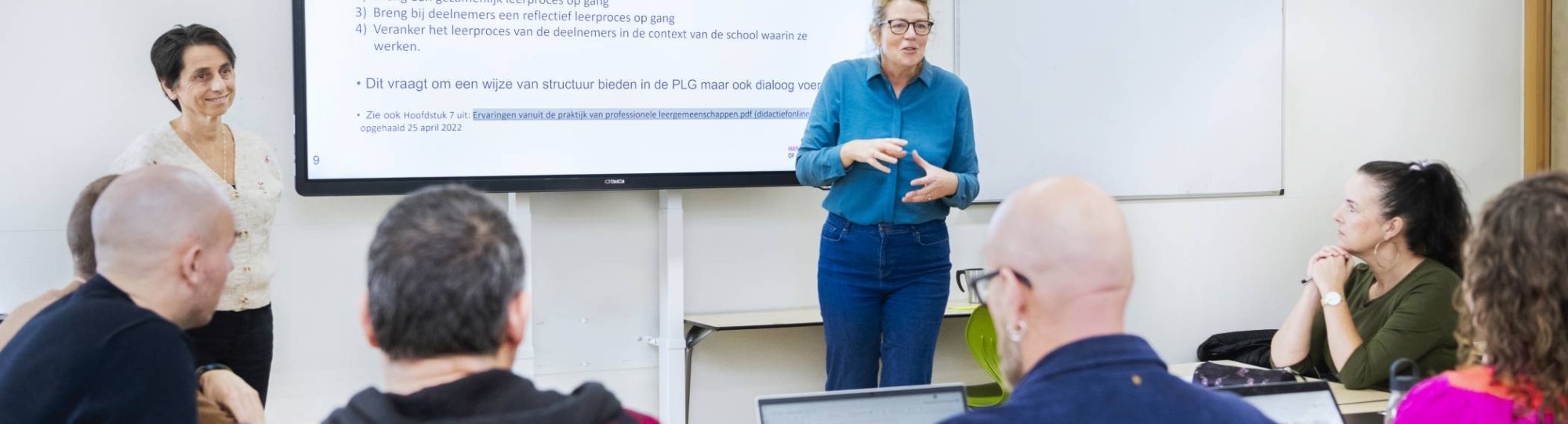 Twee vrouwelijke docenten staan enthousiast uit te leggen voor de klas, terwijl de studenten aandachtig luisteren