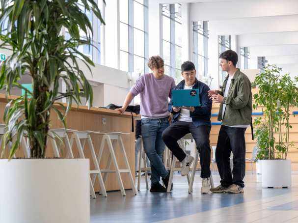 Drie jongens staan in de hal en kijken samen op een telefoon, uitgezoomde foto. 