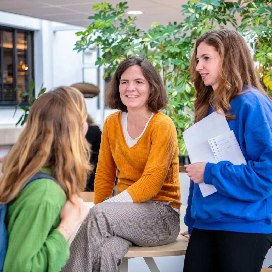 3 studenten gekleed in oranje, blauw en groen, overleggen met elkaar op de gang.