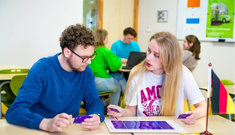 Twee studenten spelen een kaartspel met blauwe kaartjes in een gekleurd leraar Duits lokaal.
