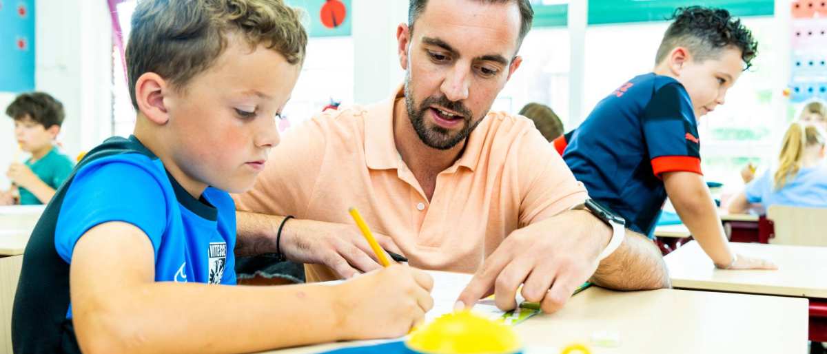 Docent van de opleiding leraar basisonderwijs, legt leerling Luc Jacobs wat uit aan tafel, terwijl Luc schrijft op een vel papier. 