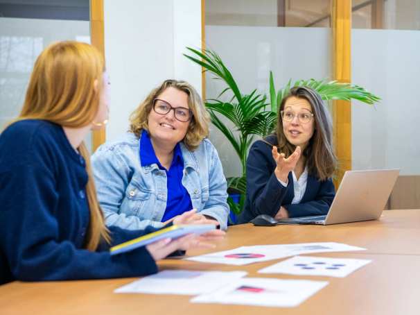 Drie mensen aan tafel in gesprek met elkaar, met een hoop papieren op tafel en een laptop