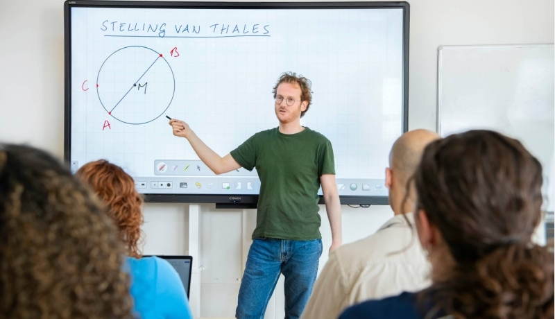 Leraar Wiskunde kijkt naar zijn klas, terwijl zijn hand wijst naar de cirkel op het bord waar 'stelling van Thales' boven staat