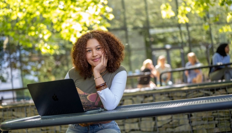 Studente kijkt in de camera, hand onder haar hoofd, werkt op laptop, buiten op bankje bij K33.