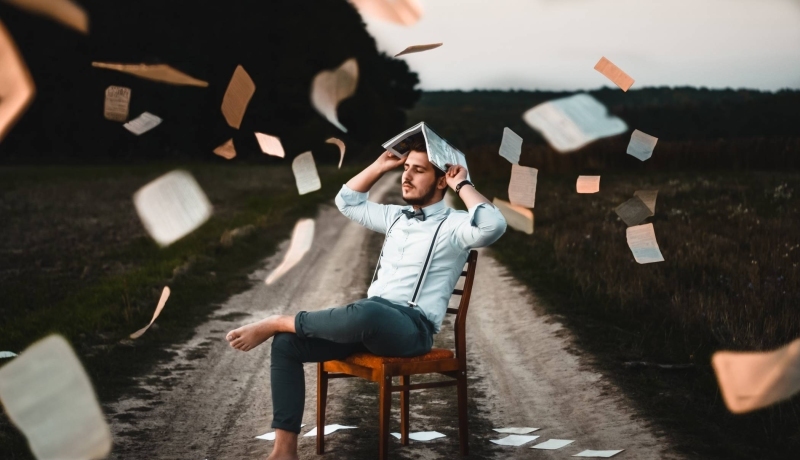 man met wit overhemd, vlinderdas en bretels zit op een stoel, midden op een zandweg. Hi houdt een boek boven zijn hoofd en losse pagina's vliegen in het rond