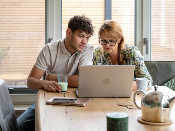 Moeder en zoon serieus bezig op de laptop, zittend aan tafel. 
