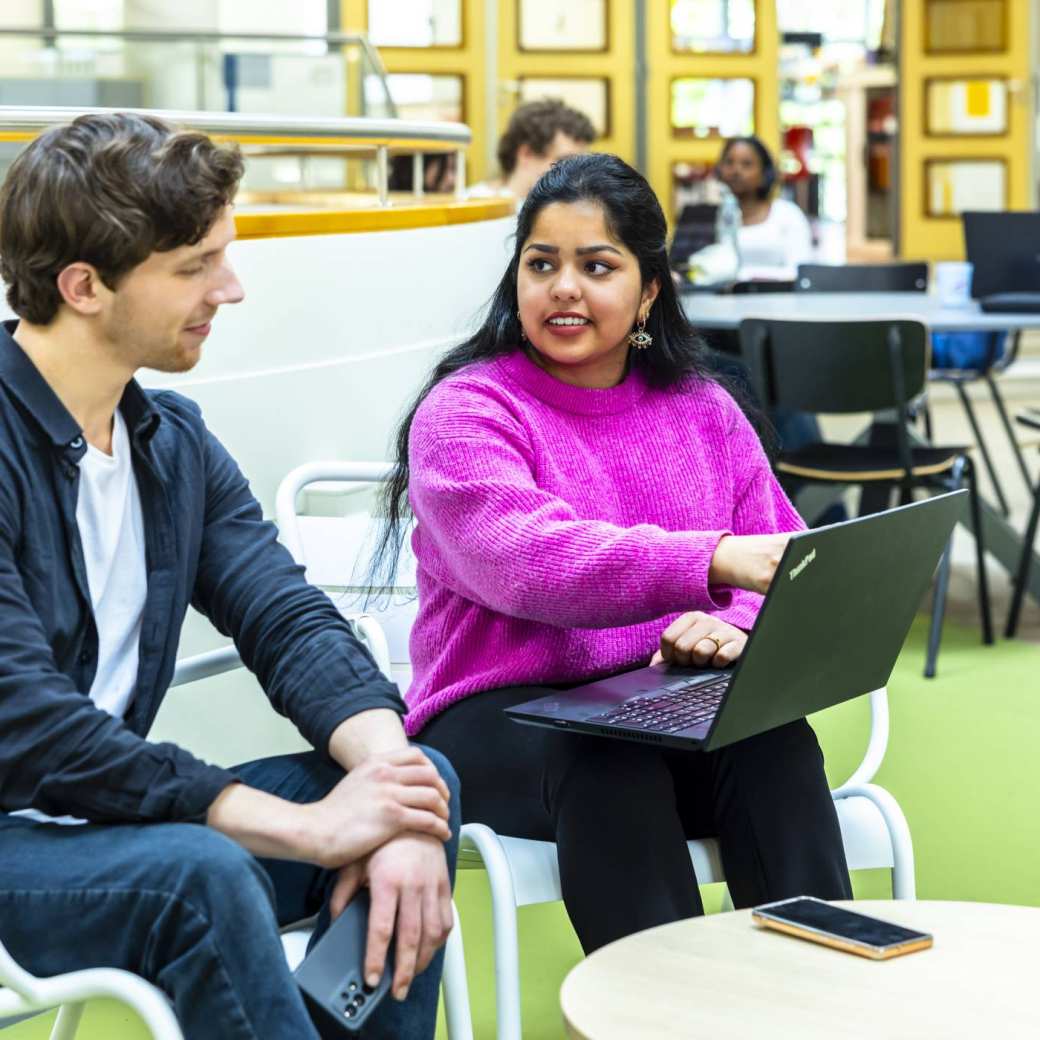 twee studenten hbo-ict zijn in gesprek terwijl een van hen een laptop op schoot heeft