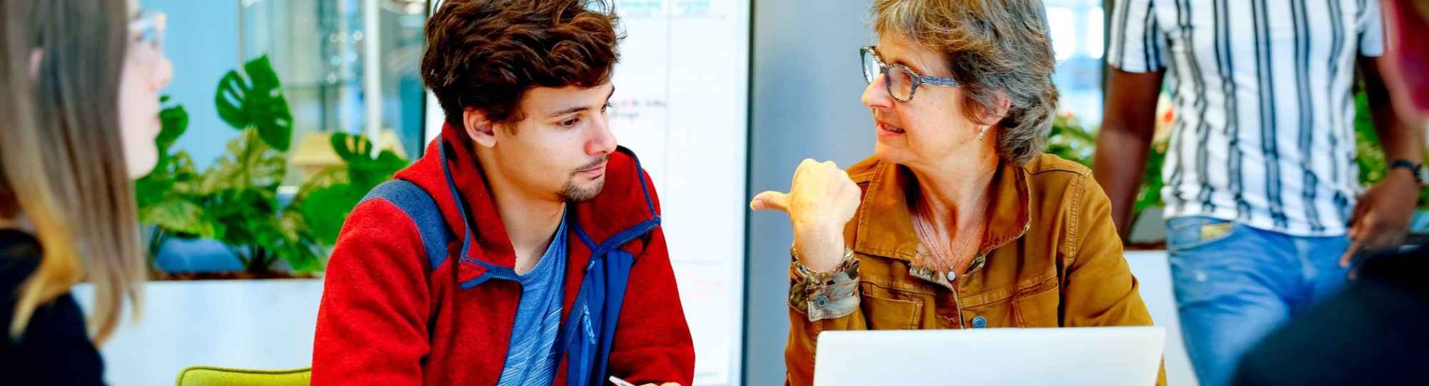 Docent zit met een laptop en drie studenten om tafel en wijst de student naast haar aan.