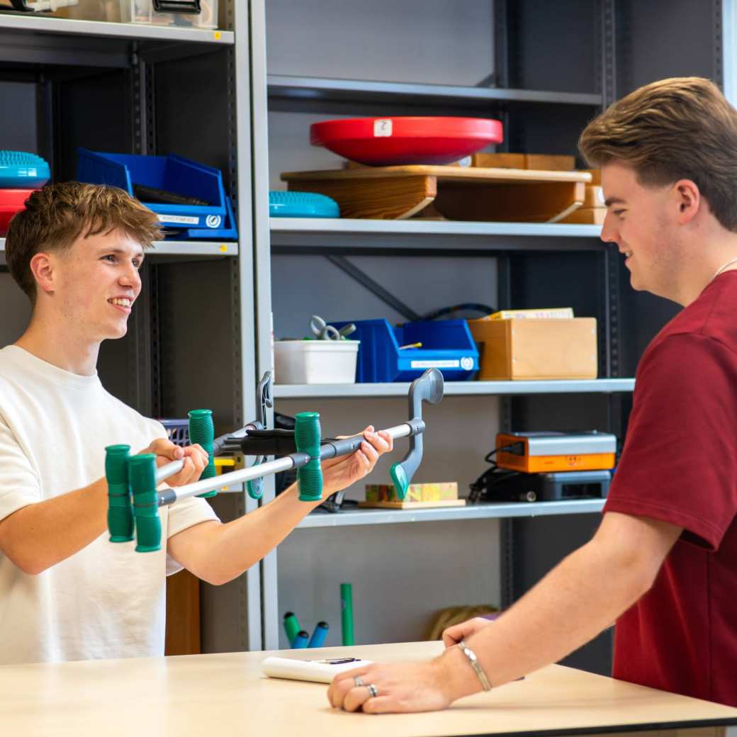 jongen laat een hulpmiddel zien aan een medestudent bij de uitleenbalie van Ergotherapie