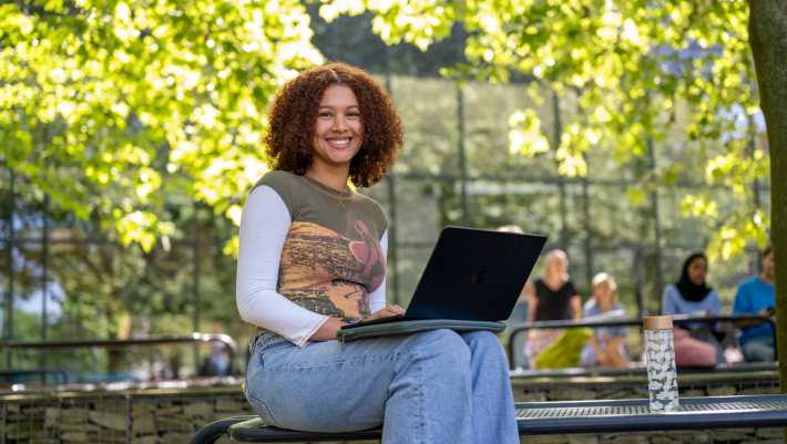 Studente kijkt in de camera, met laptop op schoot, zittend op de tafel, buiten bij K33. 