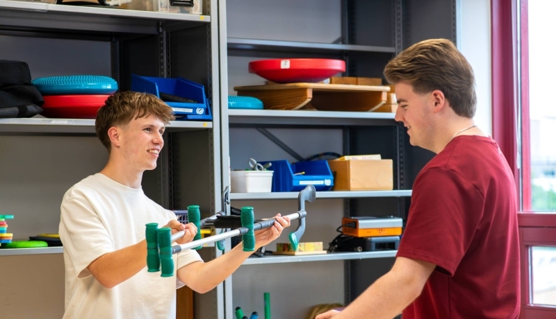 jongen laat een hulpmiddel zien aan een medestudent bij de uitleenbalie van Ergotherapie