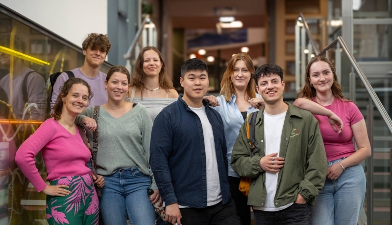 Groep studenten lachen in de camera, staan in de lichtstraat van gebouw R26. 
