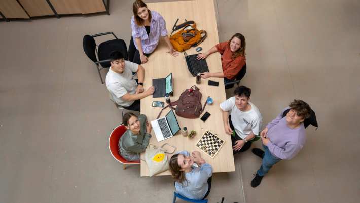 Studenten zijn aan het werk aan een tafel en kijken in camera, foto van bovenaf genomen.