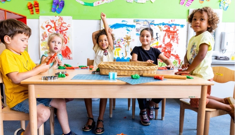 Vijf kinderen zitten aan hun tafel in de klas, waarvan één kind zijn vinger opsteekt om een vraag te stellen
