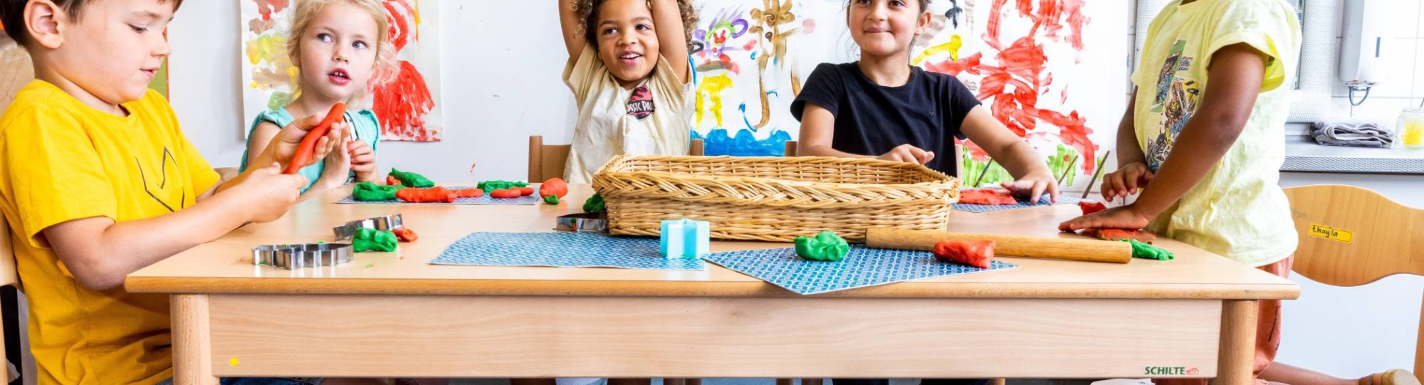 Vijf kinderen zitten aan hun tafel in de klas, waarvan één kind zijn vinger opsteekt om een vraag te stellen