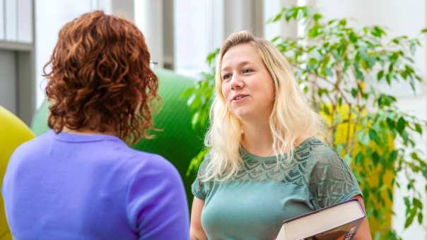 Student Leraar Wiskunde heeft boek vast en praat tegen een medestudent. Ze staan tegenover elkaar
