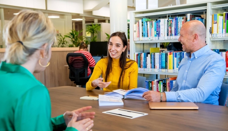 Student in overleg met twee docenten van de opleiding Bedrijfskunde deeltijd in gesprek op de HAN-campus in Arnhem, Ruitenberglaan 31. Ze lachen.