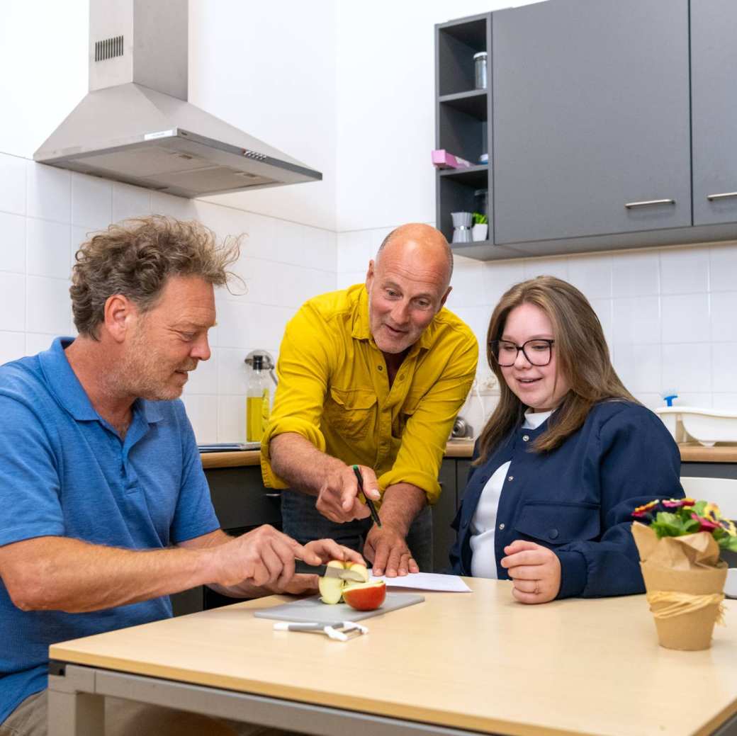 Een docent wijst naar de manier waarop een cliënt de appel snijdt en de student kijkt toe en luistert. 