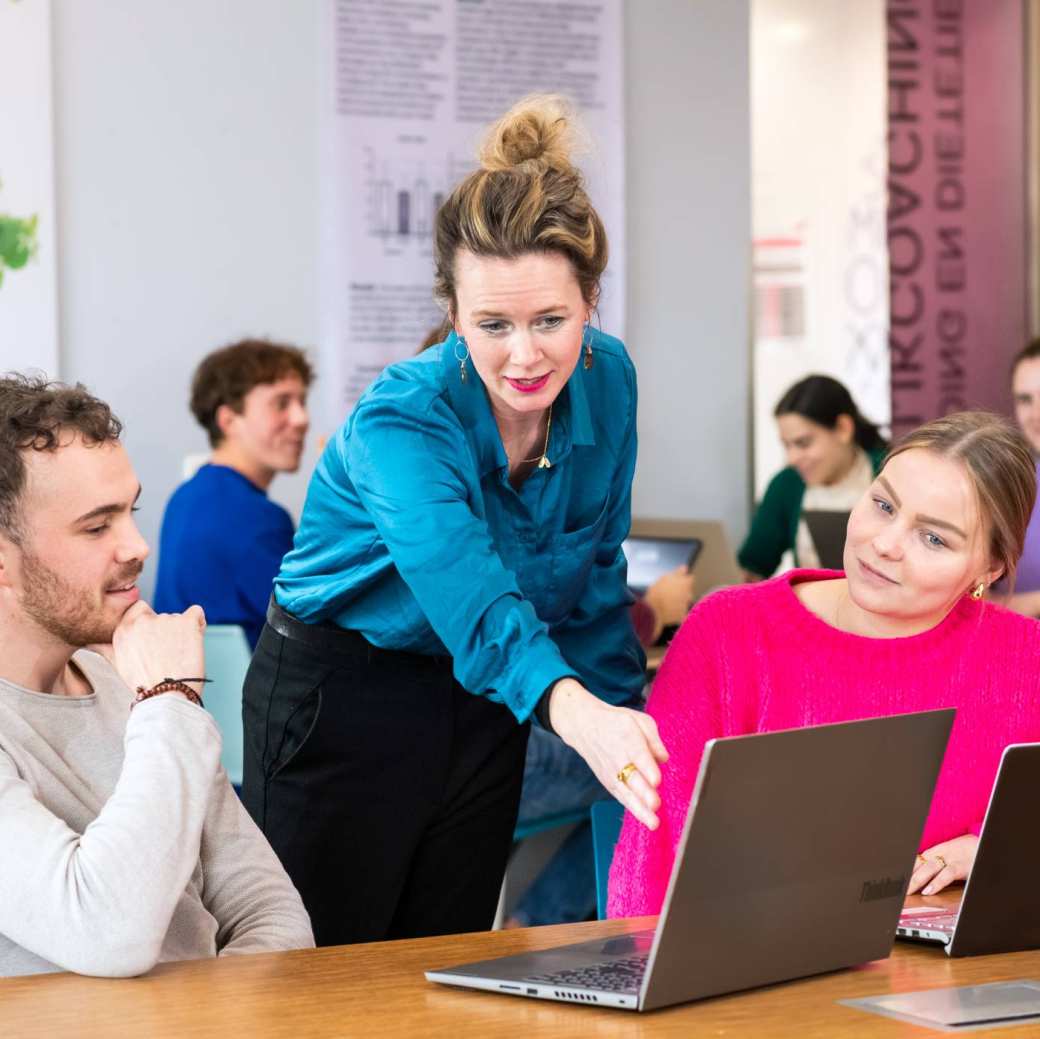 Docent helpt studenten door iets aan te wijzen op een laptop die voor de student op tafel staat. 