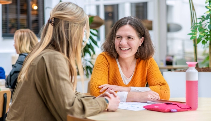 Vrouw in oranje trui lacht naar medestudent.